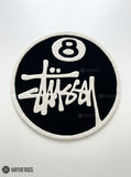 8 Ball Stüssy inspired hand-tufted custom wool rug featuring a black circular base, raised ivory 8-ball design, bold white script-style artwork, and cream border by Mayur Rugs.