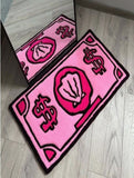 Two pink doormats with dollar sign and money bag design on a wooden floor.