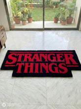 Black rug with red 'Stranger Things' text on a tiled floor, with plants outside a glass door.