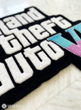 A close-up of a plush rug featuring the 'Grand Theft Auto VI' logo in white raised letters on a black background, with 'V' in light blue and pink, laid out on a white textured blanket. A 'MAYUR RUGS' watermark is visible in the bottom-left corner.