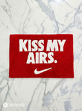 Kiss My Airs inspired hand-tufted wool rug featuring bold white typography and a swoosh-style graphic on a red background for sneakerhead and streetwear décor by Mayur Rugs.