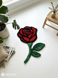 Red rose-shaped hand-tufted rug with crimson petals outlined in black, a deep green stem, and two green leaves by Mayur Rugs.