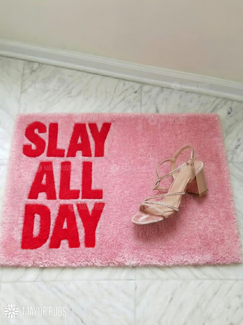 Pink doormat with 'Slay All Day' text and a sandal on a light wooden floor.