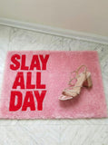 Pink doormat with 'Slay All Day' text and a sandal on a light wooden floor.