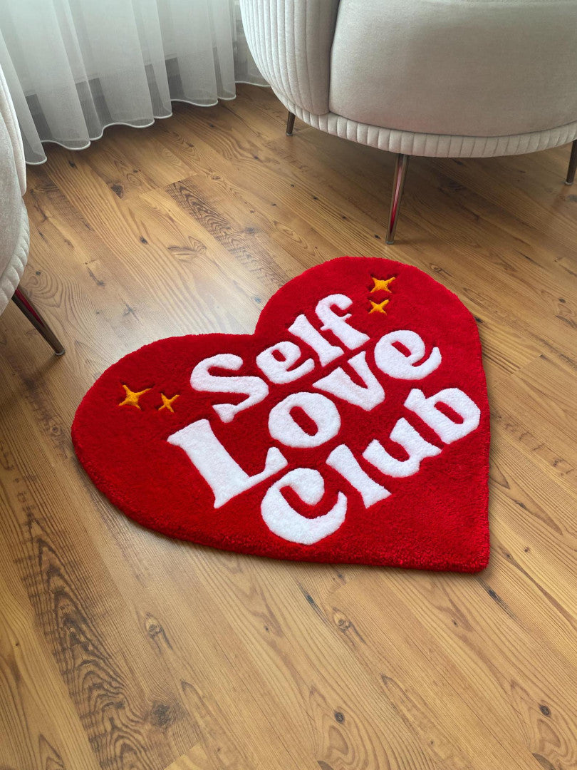 Heart-shaped red rug with 'Self Love Club' text on a wooden floor.