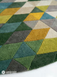 Abstract geometric round hand-tufted wool area rug featuring multicolor triangles and angular shapes in green, mustard, taupe, grey, blue, and cream by Mayur Rugs.