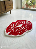 Red lips-shaped rug on a light wooden floor with a wooden cabinet and plants in the background.
