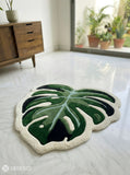 Green leaf-shaped rug on a light gray floor with a wooden cabinet and plants in the background.