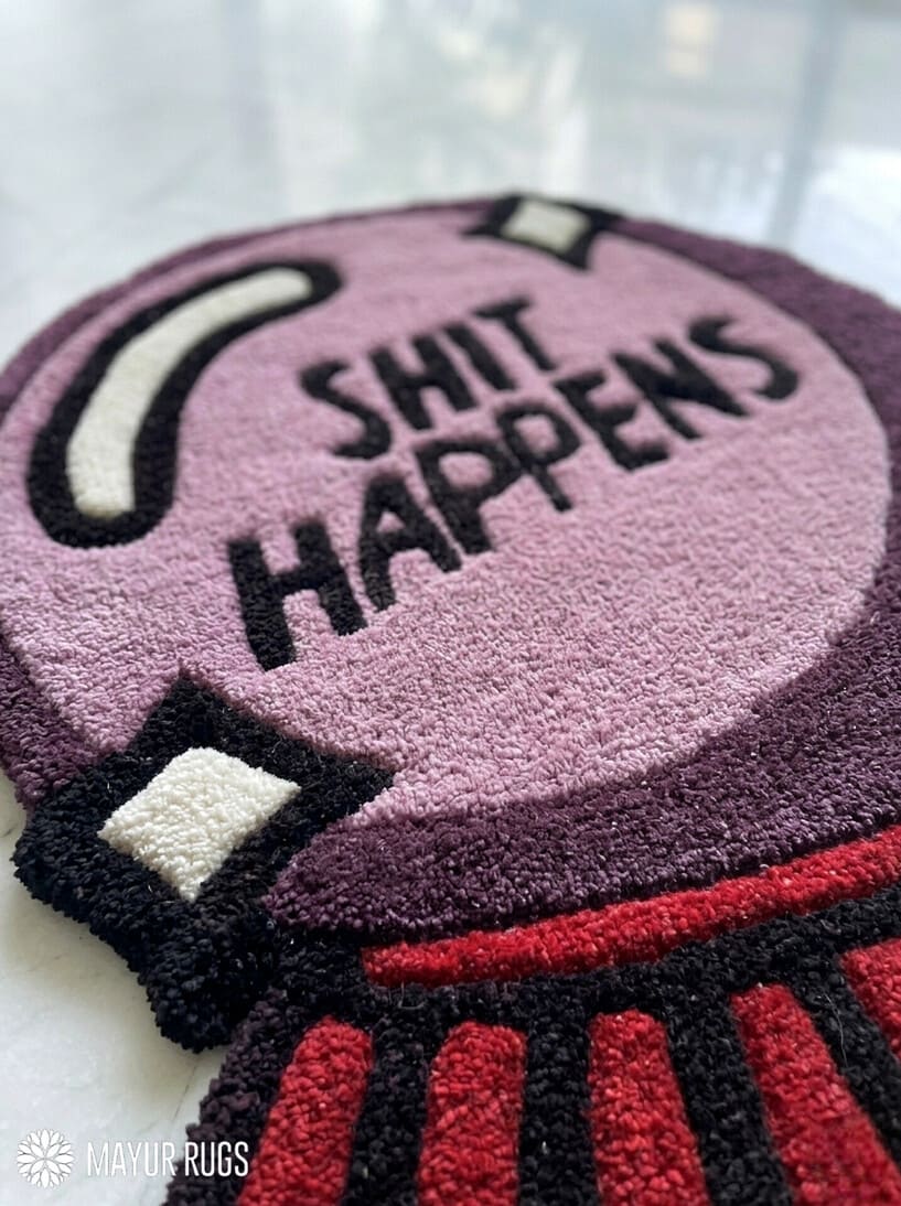 A close-up of a textured, high-pile loop rug featuring a purple and black circular design with the text 'SHIT HAPPENS' in black, accented with red and black striped details at the bottom and white details on the left.