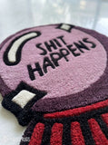 A close-up of a textured, high-pile loop rug featuring a purple and black circular design with the text 'SHIT HAPPENS' in black, accented with red and black striped details at the bottom and white details on the left.