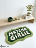 A green and white textured rug with the text 'MATCHA GIRLIE' in bold white letters on a dark green background, styled on a white floor in front of a light wood shelf with books and a plant.