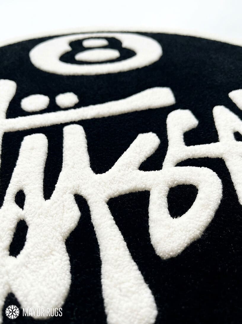 8 Ball Stüssy inspired hand-tufted custom wool rug featuring a black circular base, raised ivory 8-ball design, bold white script-style artwork, and cream border by Mayur Rugs.