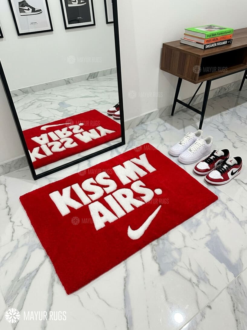 Kiss My Airs inspired hand-tufted wool rug featuring bold white typography and a swoosh-style graphic on a red background for sneakerhead and streetwear décor by Mayur Rugs.