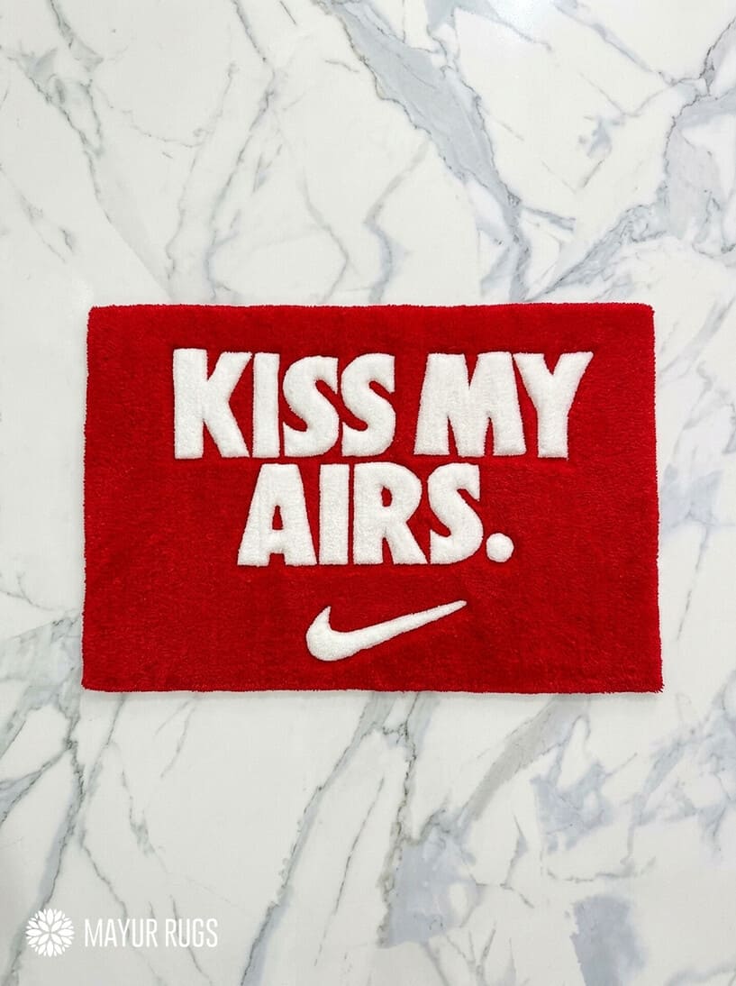 Kiss My Airs inspired hand-tufted wool rug featuring bold white typography and a swoosh-style graphic on a red background for sneakerhead and streetwear décor by Mayur Rugs.