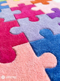 A close-up of a plush jigsaw puzzle rug with interlocking pieces in pink, blue, purple, and peach tones. A MAYUR RUGS logo watermark is visible in the bottom-left corner.