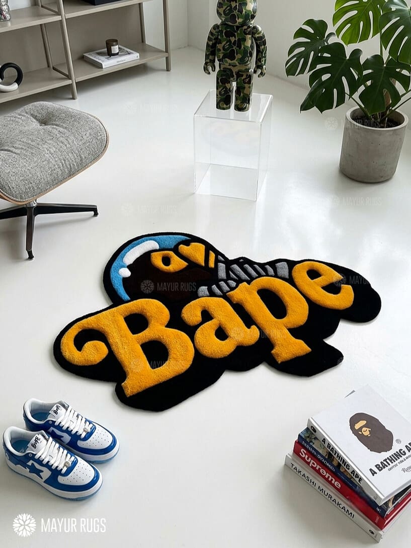 BAPE inspired ape head hand-tufted custom wool rug featuring oversized raised orange Bape typography, black graphic base, and a blue-and-white cap-wearing ape character by Mayur Rugs.