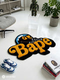 BAPE inspired ape head hand-tufted custom wool rug featuring oversized raised orange Bape typography, black graphic base, and a blue-and-white cap-wearing ape character by Mayur Rugs.