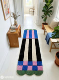 Long hand-tufted runner rug featuring cobalt blue and pink color blocks, black and ivory vertical stripes, checkerboard details, and green scalloped ends by Mayur Rugs.