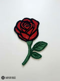 Red rose-shaped hand-tufted rug with crimson petals outlined in black, a deep green stem, and two green leaves by Mayur Rugs.